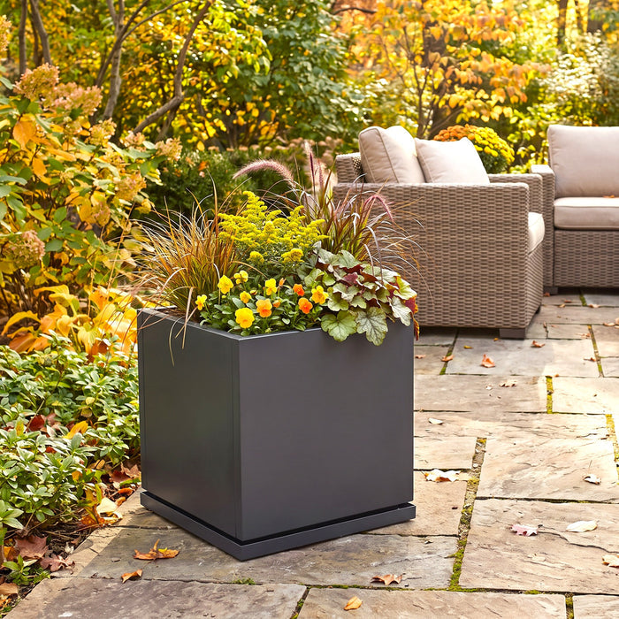 gray square planter box filled with yellow and green ornamental grasses and foliage, placed on a stone patio in a garden setting with wicker patio furniture and autumn foliage in the background.