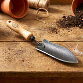 H Potter Dewit Gardening trowel with wooden handle on a wooden surface with soil and pots.